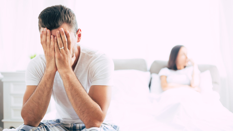 Man with head in hands at edge of bed, with woman in background looking away