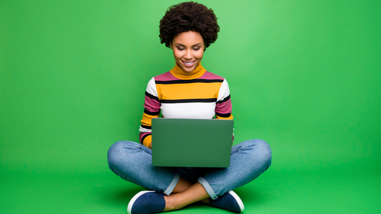 Woman sitting cross-legged typing with her laptop in her lap on a green background