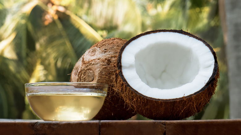 One whole and one halved coconut next to a glass bowl of coconut oil