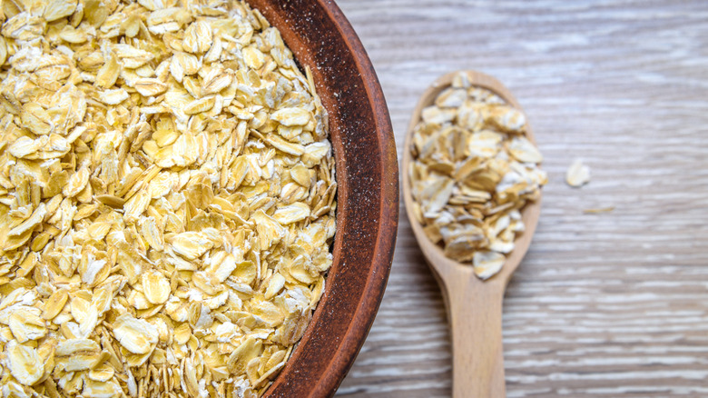 Close up of a wooden spoon filled with raw oatmeal next to a brown bowl filled with raw oatmeal