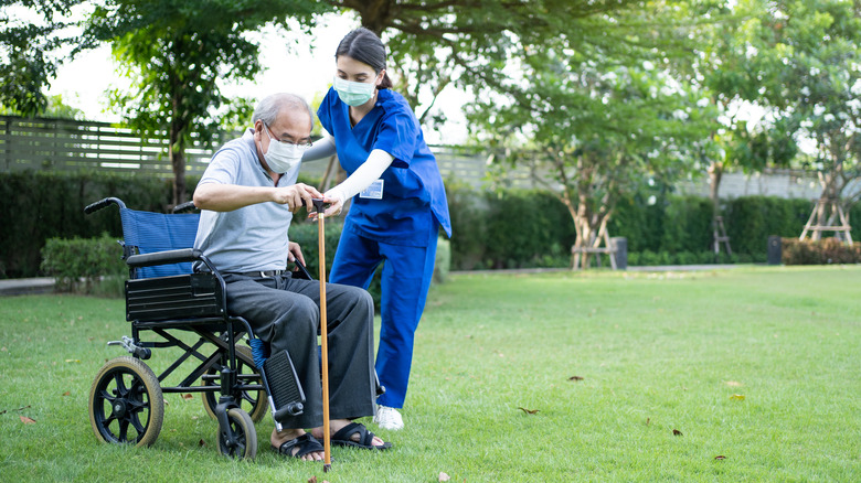 Masked doctor helping elderly older masked patient up out of wheelchair