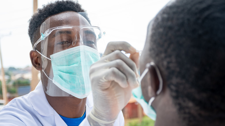 Man wearing mask, goggles, visor, and gloves using swab on patient