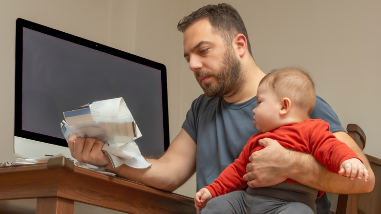 Man sitting at desk staring at pile of bills while holding baby
