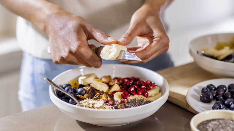 A hand slicing a banana over a bowl of fruit