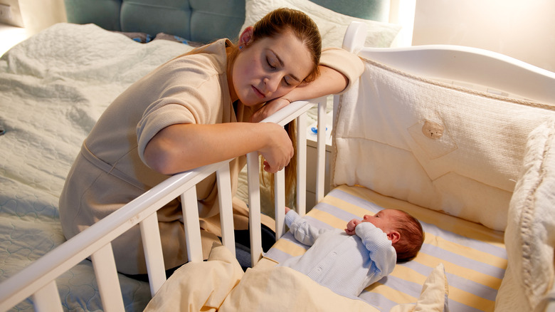 Depressed mother rests her head on baby's crib while baby sleeps in crib