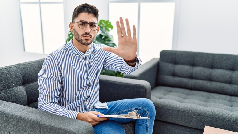 male therapist sitting on couch with a negative expression on his face, holding up hand in no sign