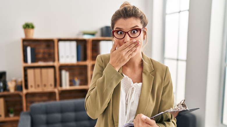 woman holding a clipboard in an office with her hand over her mouth looking shocked