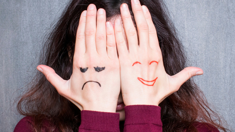 Woman holding her hands over her face, one has a smiley face and the other a sad one