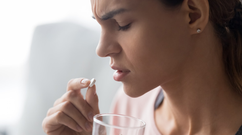 Woman in pain with closed eyes holding a white tablet pill up to her mouth along with a glass of water