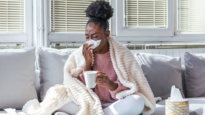 A woman on the couch with a mug while wrapped in a blanket and blowing her nose