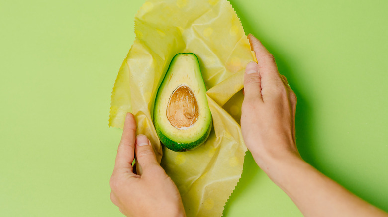 Pair of hands wrapping up a halved avocado in wax paper against green background
