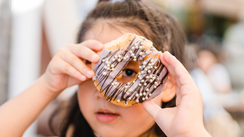 A little girl holds a donut up to her face and peers through the hole with one eye