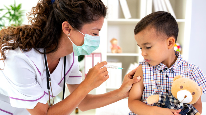 Child holding teddy bear and receiving COVID-19 vaccine shot from masked doctor