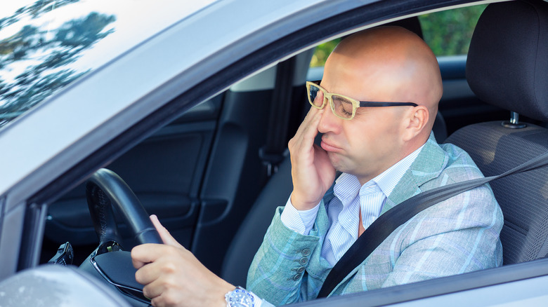 man sitting in car behind steering wheel, rubbing eye