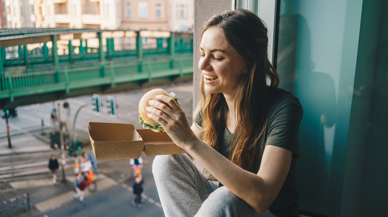 woman sitting by a window holding a fast-food sandwich