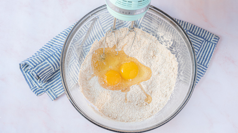 Adding eggs into dry batter in a glass mixing bowl with hand mixer