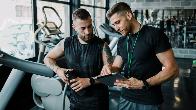 Two men talking at a gym, one holding a clipboard while pointing to it