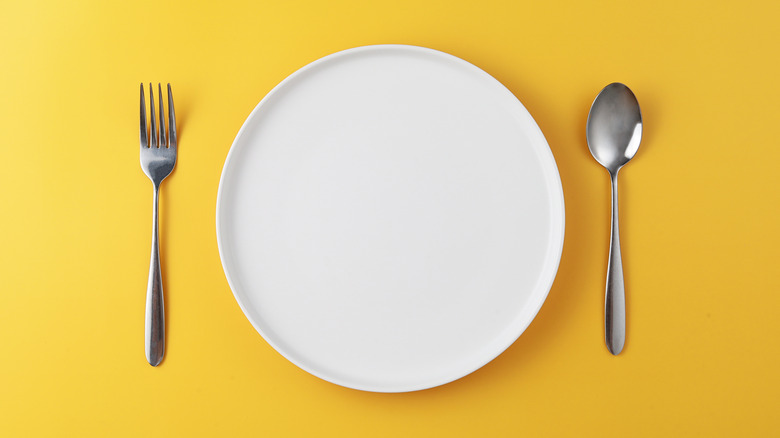 An empty white plate between a fork and a spoon against a yellow background