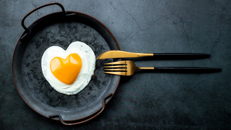 A sunny side up egg in the shape of a heart next to a knife and a fork