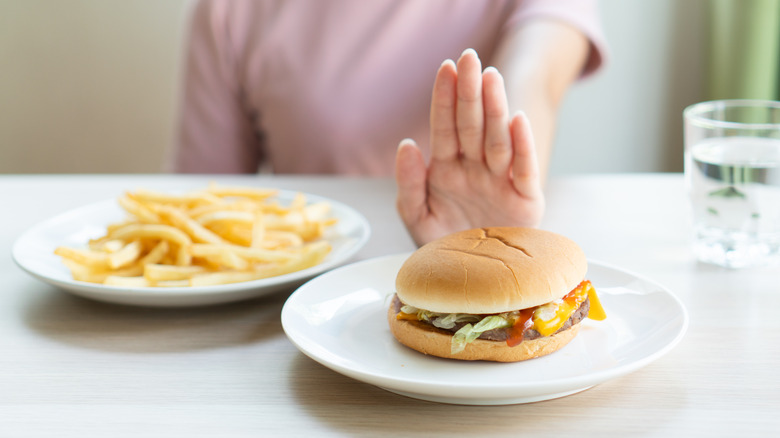 A woman pushing away a hamburger on a white plate while ignoring a plateful of fries