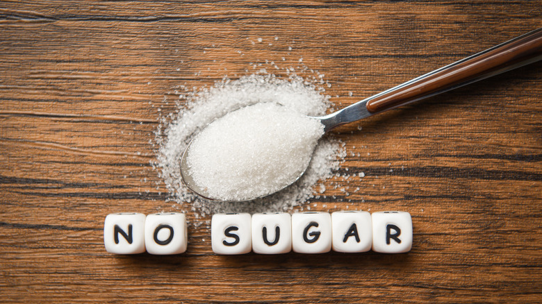 A spoonful of sugar overflowing on a wooden surface with the words "no sugar" spelled out under it