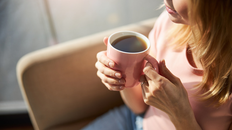 Close up on hands and torso of woman holding a pink mug of coffee while seated on the couch