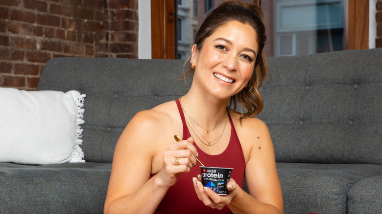 Emma sits in front of a couch, smiling and holding a yogurt