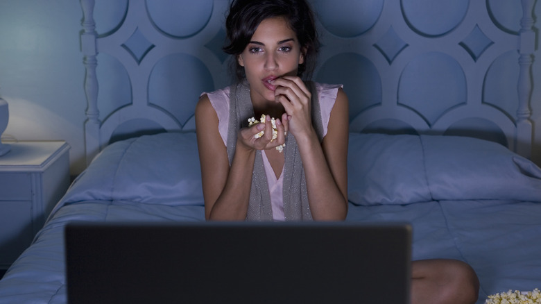 A woman eating popcorn in bed while watching a movie on her laptop