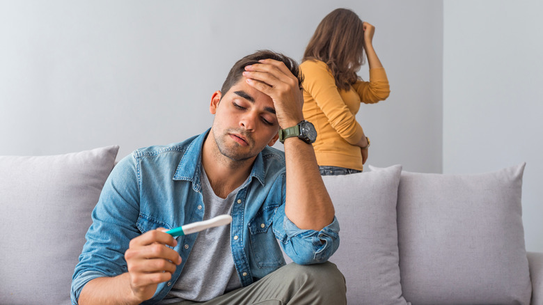 Stressed couple, man is looking at a pregnancy test and woman is standing behind the couch with her head in her hands 
