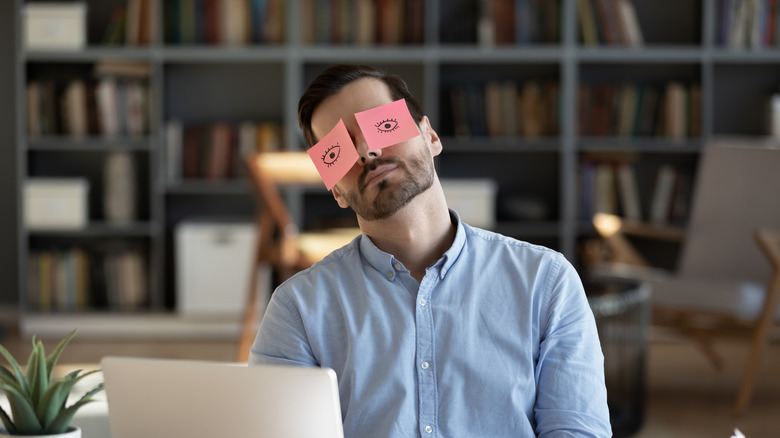 Businessman with painted eyes on sticky notes on face sleeping at workplace, sitting at desk with laptop