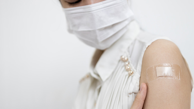 Close-up of woman with face mask holding upper arm where there is a small bandage from vaccination