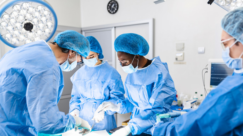 Four doctors and nurses wearing blue gowns, face masks, hair nets, and white gloves holding surgical tools