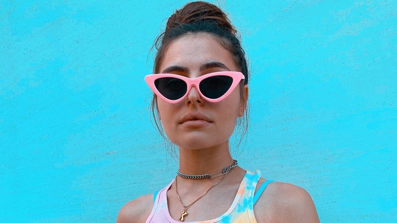 Kitty Terzo stands against a blue wall wearing pink sunglasses