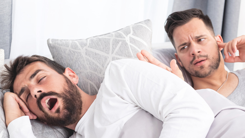 two men in bed, one with mouth open and the other plugging his ear