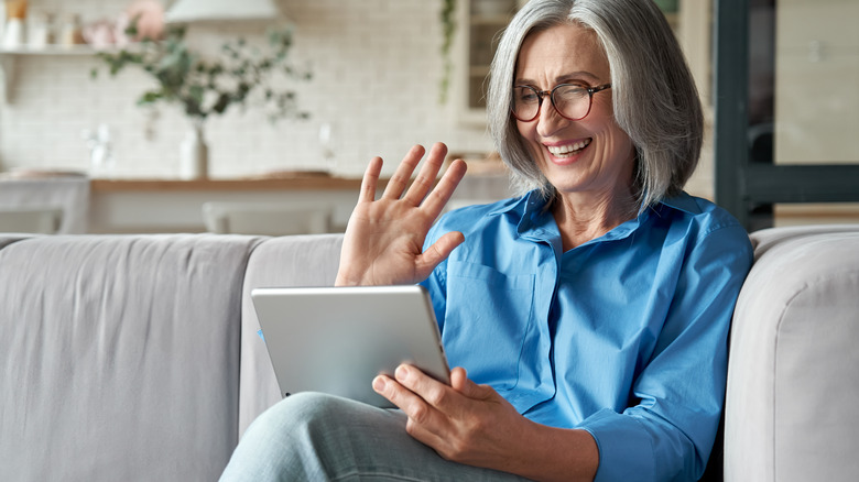 Woman sits on couch smiling and waving at her tablet, participating in telehealth appointment