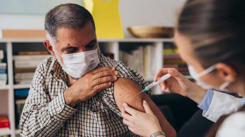 Older masked man receiving COVID-19 vaccine in upper arm from masked doctor