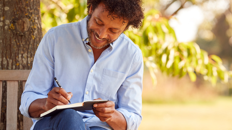 Middle-aged man smiling writing in journal while sitting outside on bench