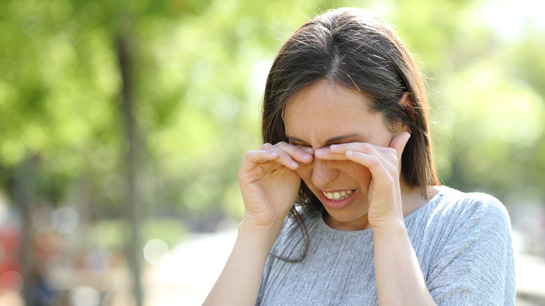 Woman in a gray shirt rubbing her eyes near some trees