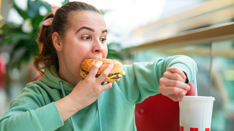 Woman in a light green hoodie looking stressed out at her watch as she eats a cheeseburger