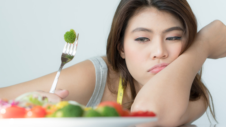 Woman looking unhappy and holding a fork with a piece of broccoli on it while staring on a plate of food