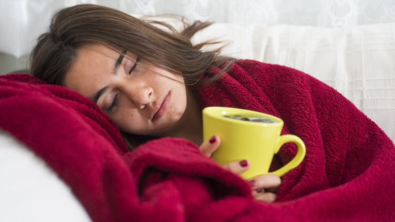 Woman wrapped up in a reddish blanket lying on her side and holding a yellow mug