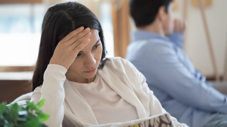 Destressed woman holding her head in the foreground while a man is out of focus in the background