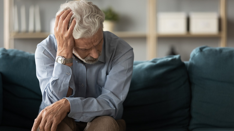 An man with white and gray hair holding his head while sitting on a couch