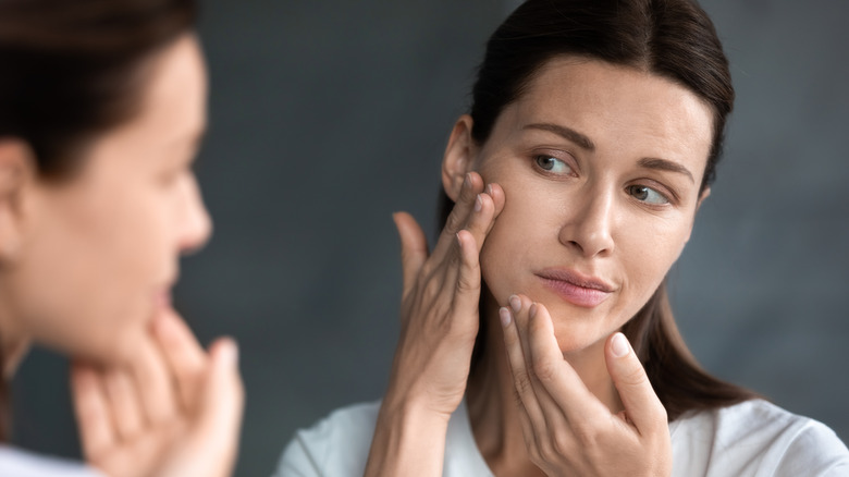 A woman with long brown hair examining the skin on her face in the mirror