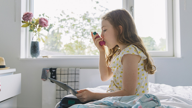 A young girl using an inhaler while sitting on her bed