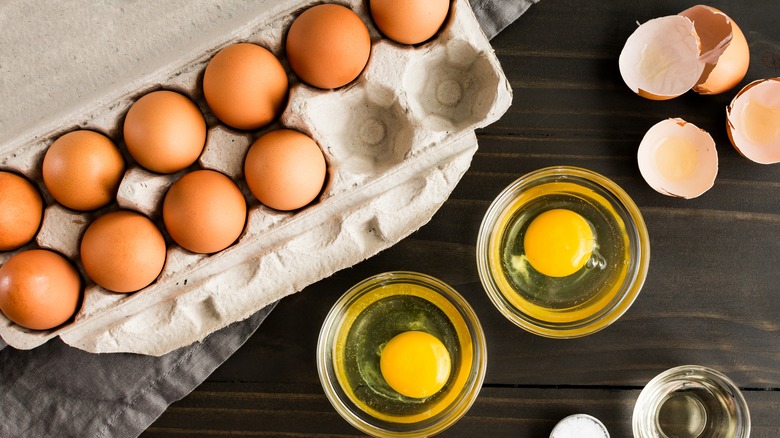 A carton of eggs next to two cracked eggs in glass bowls