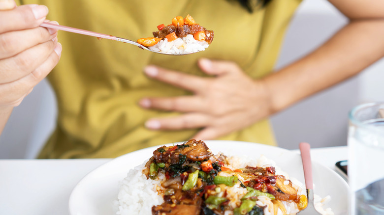 Asian woman eating spicy food and having acid reflux or heartburn hand holding a spoon with chili peppers another hand holding her stomach ache