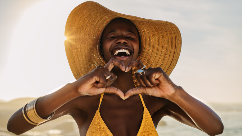 Smiling woman in bikini standing on the beach making a heart shape with her fingers.