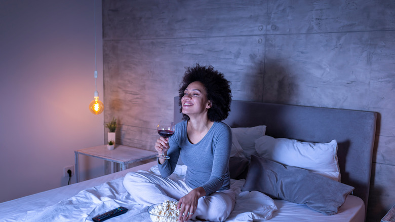 Young woman watching tv in bed with wine and popcorn happy