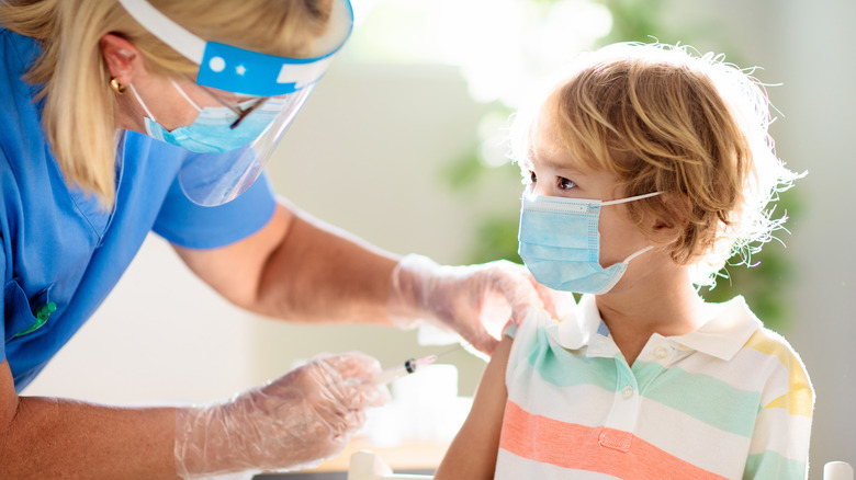 Young masked child receiving vaccine shot from masked nurse with face shield